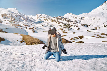 Full body of happy funny boy in outwear sitting on snow and exploring virtual reality on sunny winter day in mountains