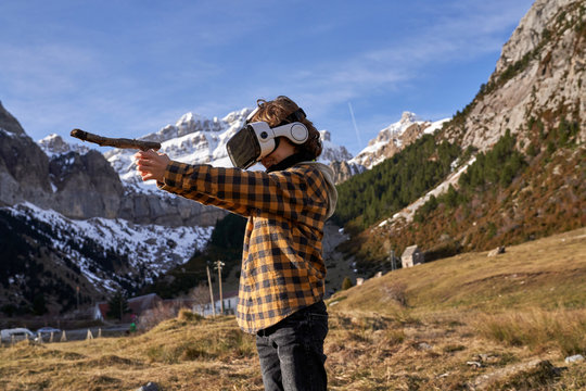 Active smart boy looking away in VR glasses playing with stick standing on stone in mountain valley