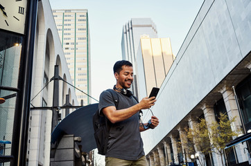 Contemporary Hispanic male tourist in casual wear and sunglasses with backpack and headphones using mobile phone at modern city street