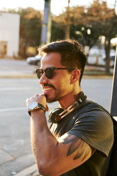 Young Hispanic man in casual wear and stylish sunglasses with headphones and backpack looking away at city street