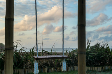 A  wooden swing with the view on Nyang Nyang Beach. The swing has very simple construction. There is green grass around the swing. Hidden getaway. Collecting happy moments.