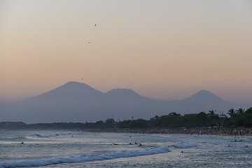 Golden hour on Seminyak Beach, Bali, Indonesia. The sky has pinkish color. The beach bay is washed by gentle waves, There are high volcanic mountains in the back. Idyllic and relaxing moment