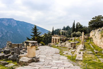 Fototapeta na wymiar Rock walkway down hill in ancient Delphi Greece past ionic columns and parts of temples and past a reconstructed treasury with mountain in distance