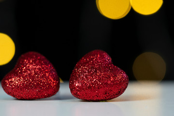 two red shiny hearts lie on a white table. Black wall with bokeh. Garlands. Valentine's day.