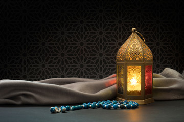 Ramadan Lantern and Blue rosary beads in front of dark arabesque background.