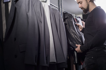 Portrait of a young bearded millennial guy. In a men's clothing store, a guy in the middle of a counter with business jackets selects a leather brutal rock and roll jacket