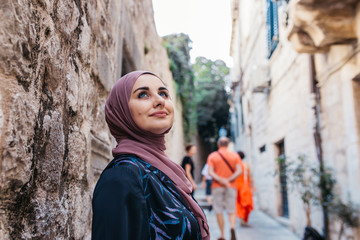 Portrait of beautiful woman with hijab. She is happy and relaxed, while tourist passing by her in narrow old street of Dubrovnik. 2018.