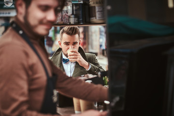 Stylish and elegant young male customer sitting outdoors next to coffee making barista in a mobile coffee shop in a city emporium, wearing green wool coat, white shirt and bow tie, taking a sip of