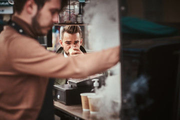 Hipster and elegant young male customer sitting outdoors next to coffee making barista in a mobile coffee shop in a city emporium, wearing green wool coat, white shirt and bow tie, taking a sip of