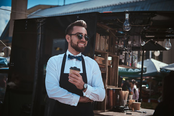 Handsome looking man barista working in a mobile coffee shop outdoors in the city emporium wearing apron and white shirt with bow tie, - obrazy, fototapety, plakaty