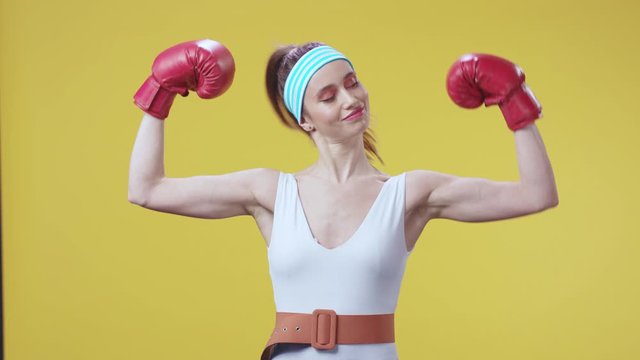 Happy strong caucasian woman fitness coach showing her biceps and testing arm musles smiling proud on camera posing and training at yellow wall background.