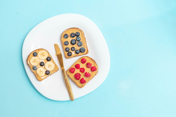 Peanut butter toast snack with blueberry, raspberry, banana on white plate on light blue background. Top view, copy space