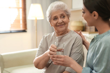 Young woman giving water to elderly lady indoors. Senior people care
