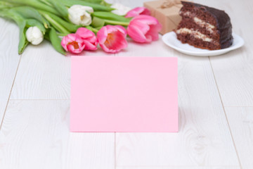 Paper card, bouquet of pink tulips, chocolate cake. pressent box on white wooden background. Copy space, close up. Mother's, women's day concept.