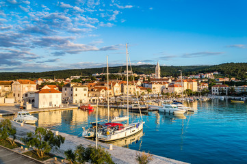 Supetar on Brac Island near Split, Croatia. Small seaside town with promenade and harbor with white boats, palm trees, cafes, houses and church. Tourists walk the street on sunny day at sunset