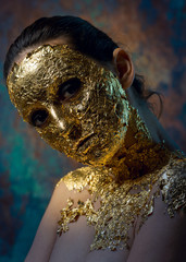 Girl with a mask on her face made of gold leaf. Gloomy studio portrait of a brunette on an abstract background.