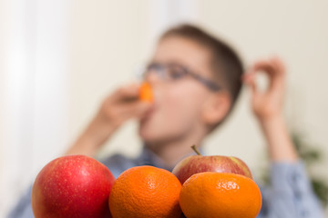 Mandarynki i jabłka na pierwszym planie. Na drugim planie chłopiec który zjada ze smakiem owoce.
