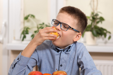 Chłopiec w niebieskiej koszuli i okularach siedzi przy stole i trzyma w dłoni jabłko które je ze smakiem. - obrazy, fototapety, plakaty