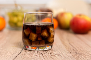 Szklanka wypełniona kostkami cukru zalana colą stoi na kuchennym blacie. W tle na blacie leżą owoce jabłka, mandarynki, winogrona.   