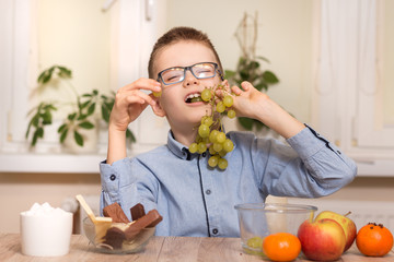 Uśmiechnięty chłopiec siedzi przy stole i zjada owoce. Trzyma w zębach owoce winogrona. - obrazy, fototapety, plakaty