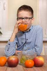 Uśmiechnięty chłopiec siedzi przy stole i pije ze szklanki sok. Owoce mandarynki, jabłka i winogrona leżą na stole. - obrazy, fototapety, plakaty