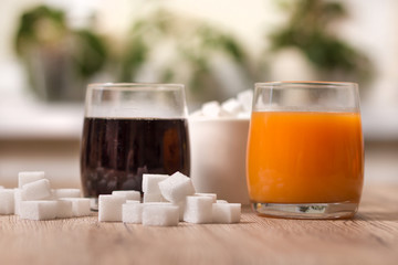 Kostki cukru porozrzucane leżą na blacie kuchennym. W jednej szklance stoi cola a w drugiej sok. Na drugim planie cukiernica wypełniona kostkami cukru. - obrazy, fototapety, plakaty