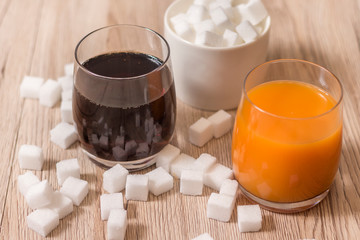 Kostki cukru porozrzucane leżą na blacie kuchennym. W jednej szklance stoi cola a w drugiej sok. Na drugim planie cukiernica wypełniona kostkami cukru.