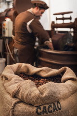 Jute bag full of cocoa beans in a chocolate maker workshop, with a male chocolatier working on conching and melanger equipment on the background - 324074696