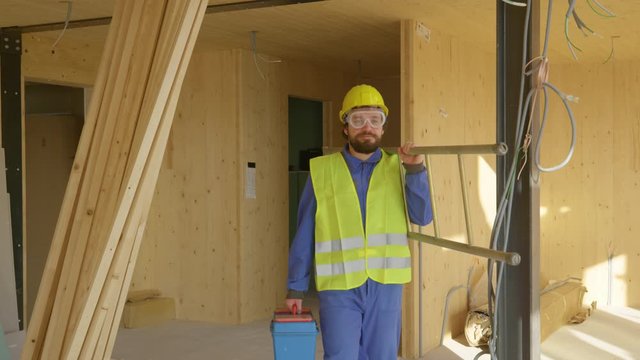 SLOW MOTION, CLOSE UP: Worker coming to work carries his metal toolbox and ladder across the room of a prefabricated house. Builder leaves the construction site with his toolbox and ladder in hand.