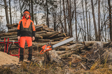 A chainsaw operator is next to a tree trunk and a big orange chainsaw. Protective equipment is...
