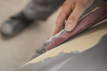 A hand of a man is seen sanding an old vintage car using a block of wood and brown sand grit paper....