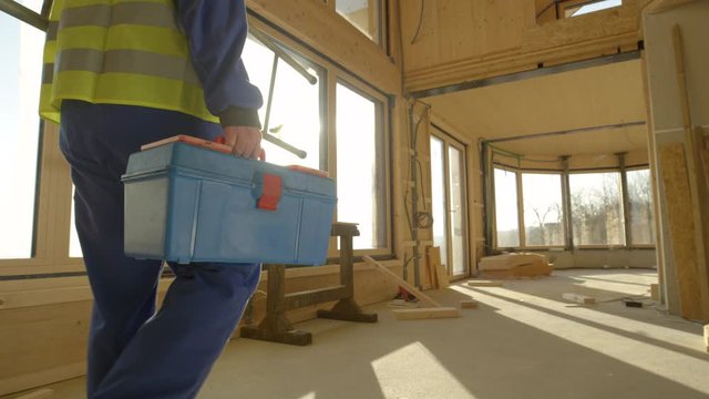 SLOW MOTION, LENS FLARE, CLOSE UP: Unrecognizable builder arrives to work carrying his toolbox and ladder. Golden sunbeams shine on worker coming to work on a contemporary prefabricated wooden house.