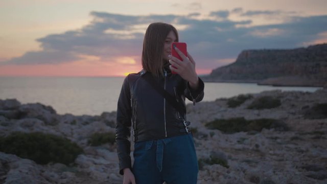 Happy esctatic Caucasian woman making cute selfies on modern smartphone under sunset in spectacular landscape with calm blue ocean.