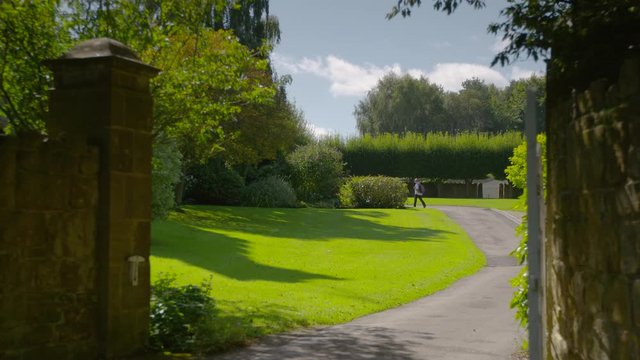 Hiker with backpack walking along a paved path through a nicely landscaped garden, passing trees, shrubs, and a green manicured lawn