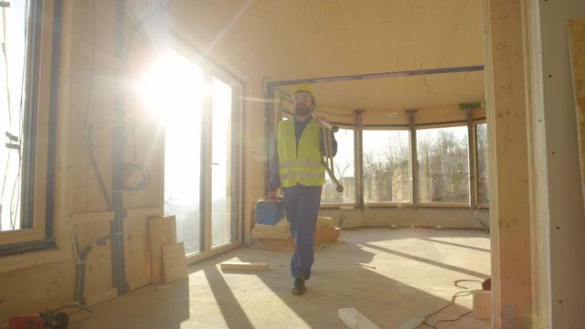 SLOW MOTION, CLOSE UP, LENS FLARE: Caucasian worker wearing blue overalls and a yellow hard hat arrives to work on a sunny morning. Contractor carries his toolbox and ladder to work on a sunny day.