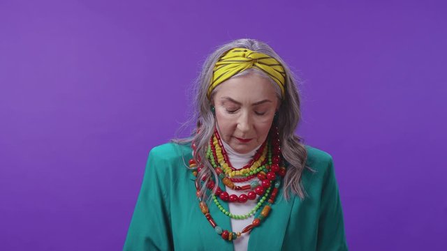 Attractive old caucasian woman in fashion looks down then smiles cheerfully at camera against purble background. Stylish grandma. Copy space.