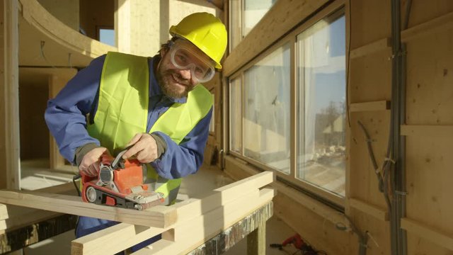 SLOW MOTION, PORTRAIT, CLOSE UP: Smiling builder working on a prefabricated house buffs a long wooden beam. Cheerful Caucasian contractor uses an electric sander to smooth out the edge of a plank.