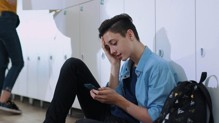 Profile of upset college student sitting by lockers in the school corridor. handsome young boy in trouble using a smartphone trying to get things done.