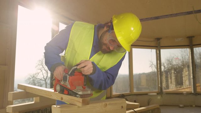 SLOW MOTION, LENS FLARE, CLOSE UP: Bearded Caucasian contractor diligently smoothens a wooden beam with an electric sander. Young worker working in a glued laminated timber house buffs a wooden board