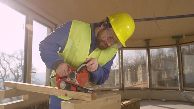 SLOW MOTION, LENS FLARE, CLOSE UP: Bearded Caucasian contractor diligently sands a wooden beam with an electric sander. Young worker working in a glued laminated timber house buffs a wooden board
