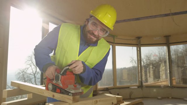 SLOW MOTION, LENS FLARE, PORTRAIT: Content contractor building a modern cross-laminated house smoothens a wood board. Caucasian construction worker smiles while sanding a wooden beam on sunny day.