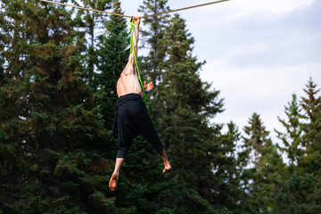 A close up view of a strong man hanging by one hand from a tightrope wire during a gymnastic routine. in woodland at a festival celebrating earth