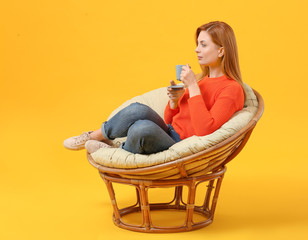 Beautiful woman drinking coffee while sitting in armchair against color background