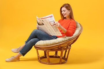 Beautiful woman reading newspaper while sitting in armchair against color background