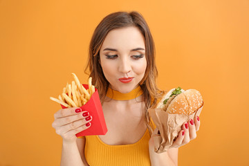 Portrait of beautiful young woman with burger and french fries on color background