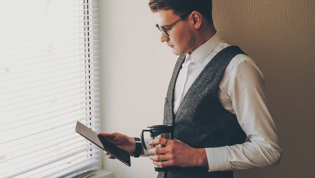 A busy caucasian businessman dressed in a suit is holding a tablet near the window while drinking a coffee - Powered by Adobe