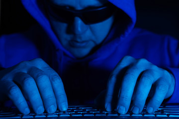 A man in glasses and a blue hoodie works at a computer. Soft focus