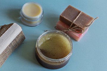 Spa cosmetics, flower soap, handmade sugar scrub with coconut oil in a glass jar, face cream and washcloth made of natural material on a blue background