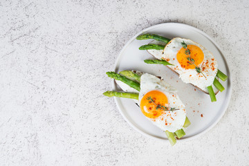 Crispy toast with cream cheese cream, fried egg and asparagus in a plate on a white background.