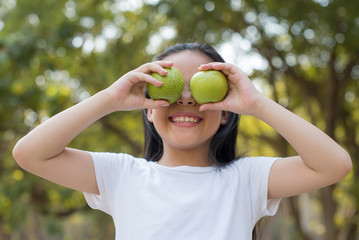 photo happy Little asian girl child standing with big smile. holding green apple in your hand.fresh healthy green bio background with abstract blurred foliage and bright summer sunlight